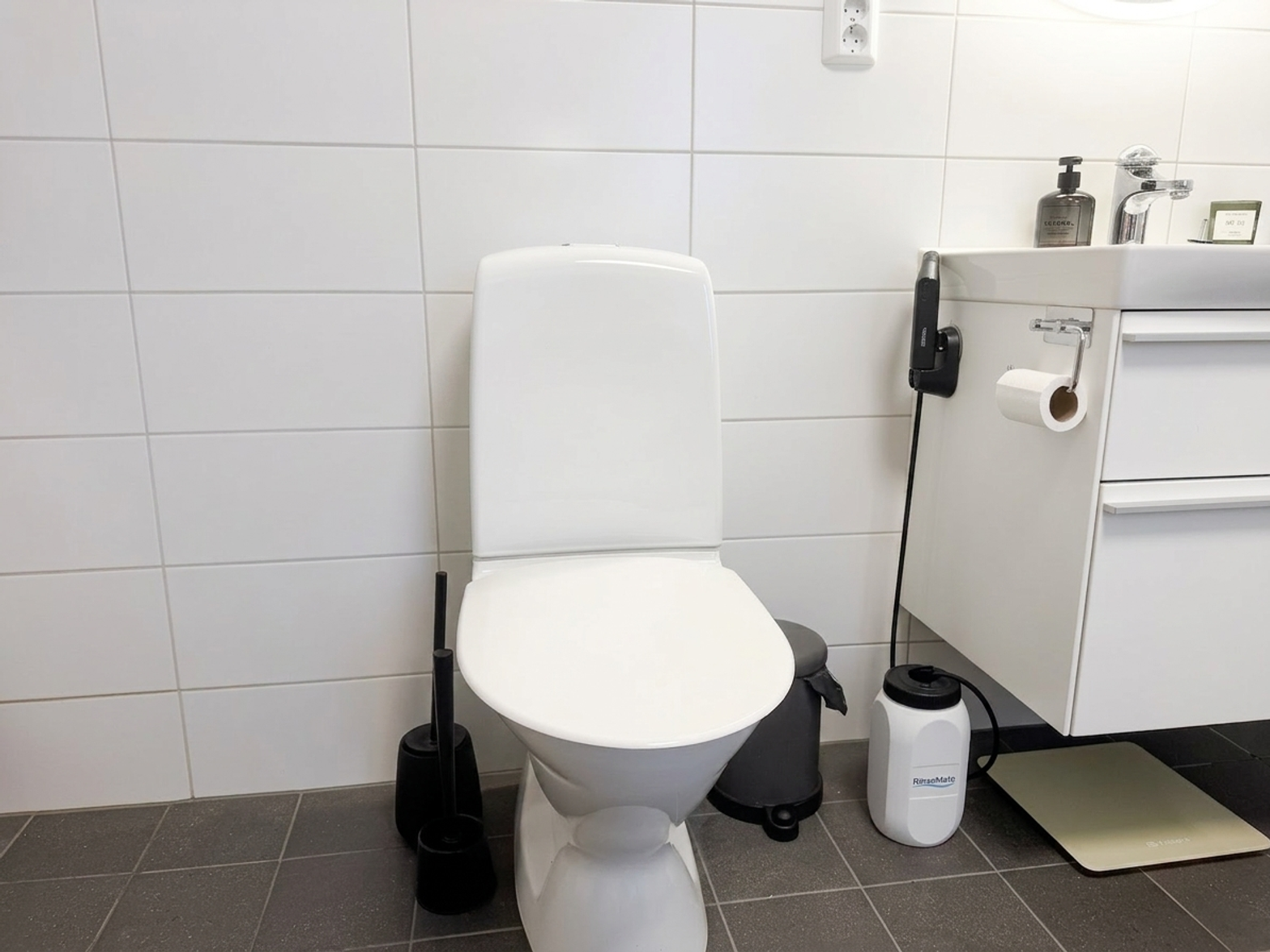 A RinseMate portable bidet with a 2.5-litre floor tank connected by a hose to a handheld wand, standing beside a white toilet in a tiled bathroom.